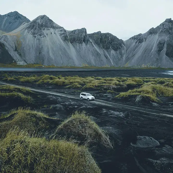 A white SUV driving on a rugged road through a dramatic black volcanic landscape with steep, rocky mountains in the background.