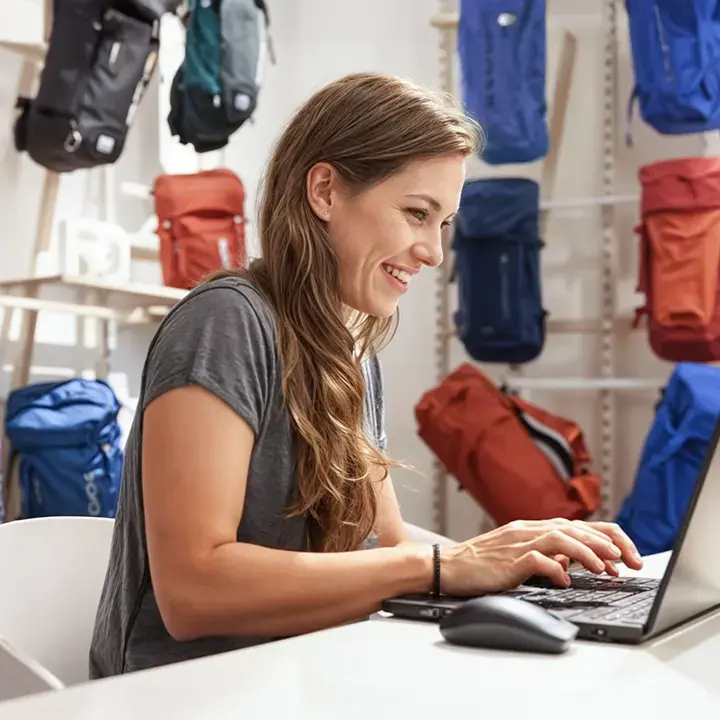 Smiling woman working on a laptop in a showroom with various backpacks displayed on shelves in the background.