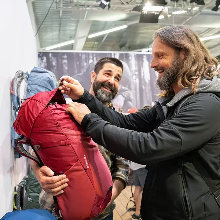 Two men smiling and examining a red backpack at an indoor event or exhibition, with other backpacks displayed in the background.