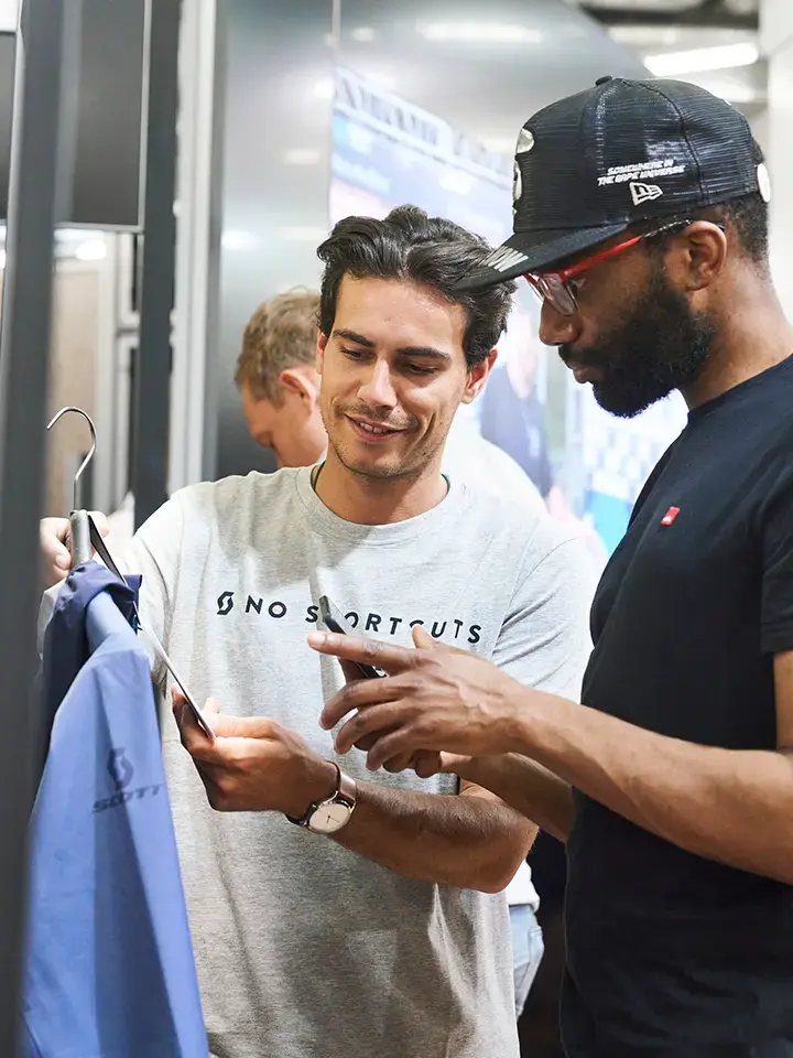 Two men discussing a product at an exhibition, one holding a jacket on a hanger and the other looking at a phone.