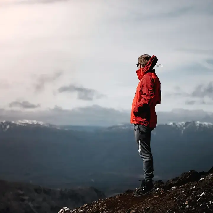 Man in a red jacket standing on a mountain top, gazing at the expansive landscape and cloudy sky in the distance.