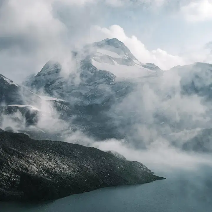 Snow-covered mountain peak partially shrouded in mist, with a calm lake in the foreground under a cloudy sky.