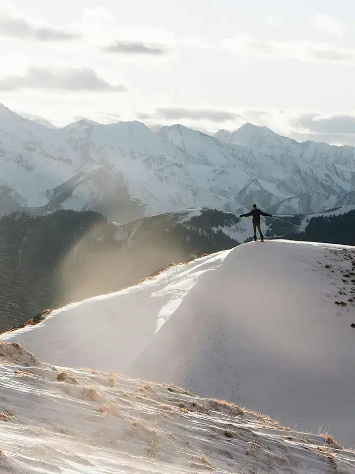 Person standing on a snowy mountain ridge, holding ski poles and looking out at a breathtaking view of snow-covered peaks under a cloudy sky.