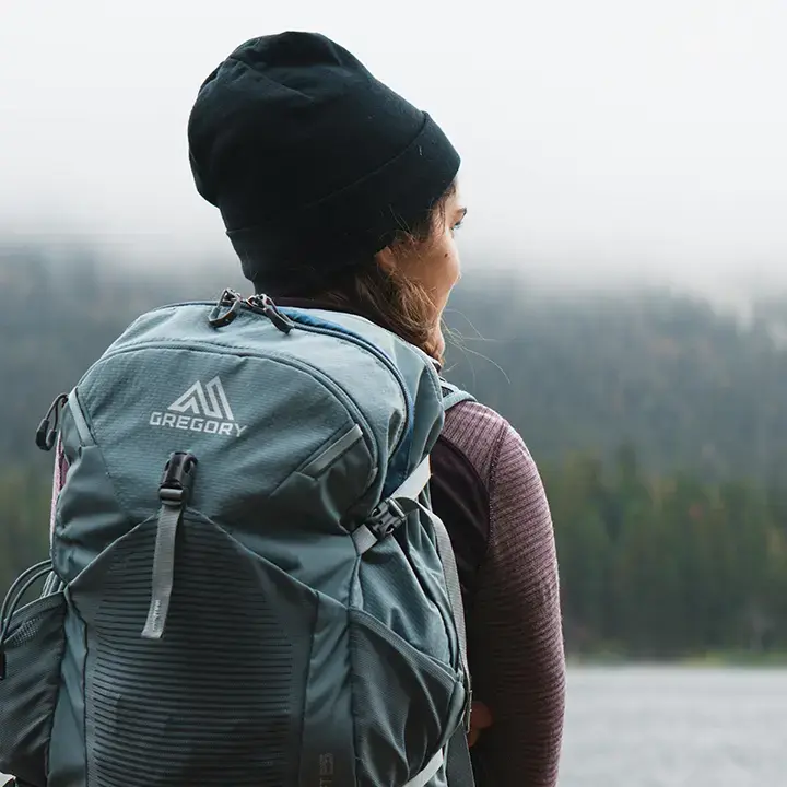 Back view of a woman wearing a beanie and a Gregory backpack, looking out at a foggy landscape near a lake.