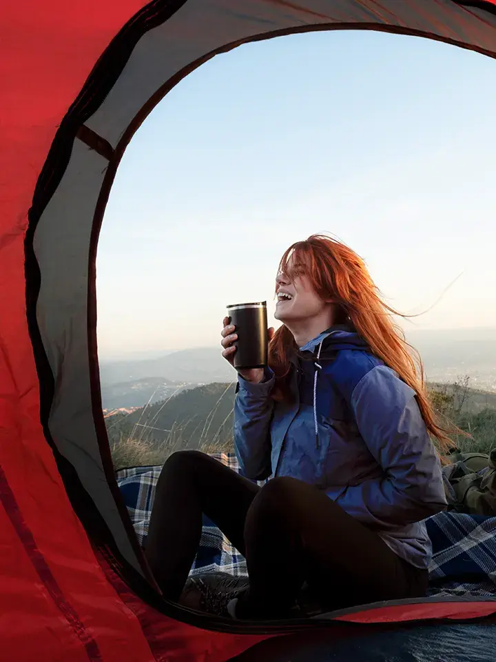 Smiling woman sitting at the entrance of a red tent, enjoying a drink with a scenic mountain view in the background.